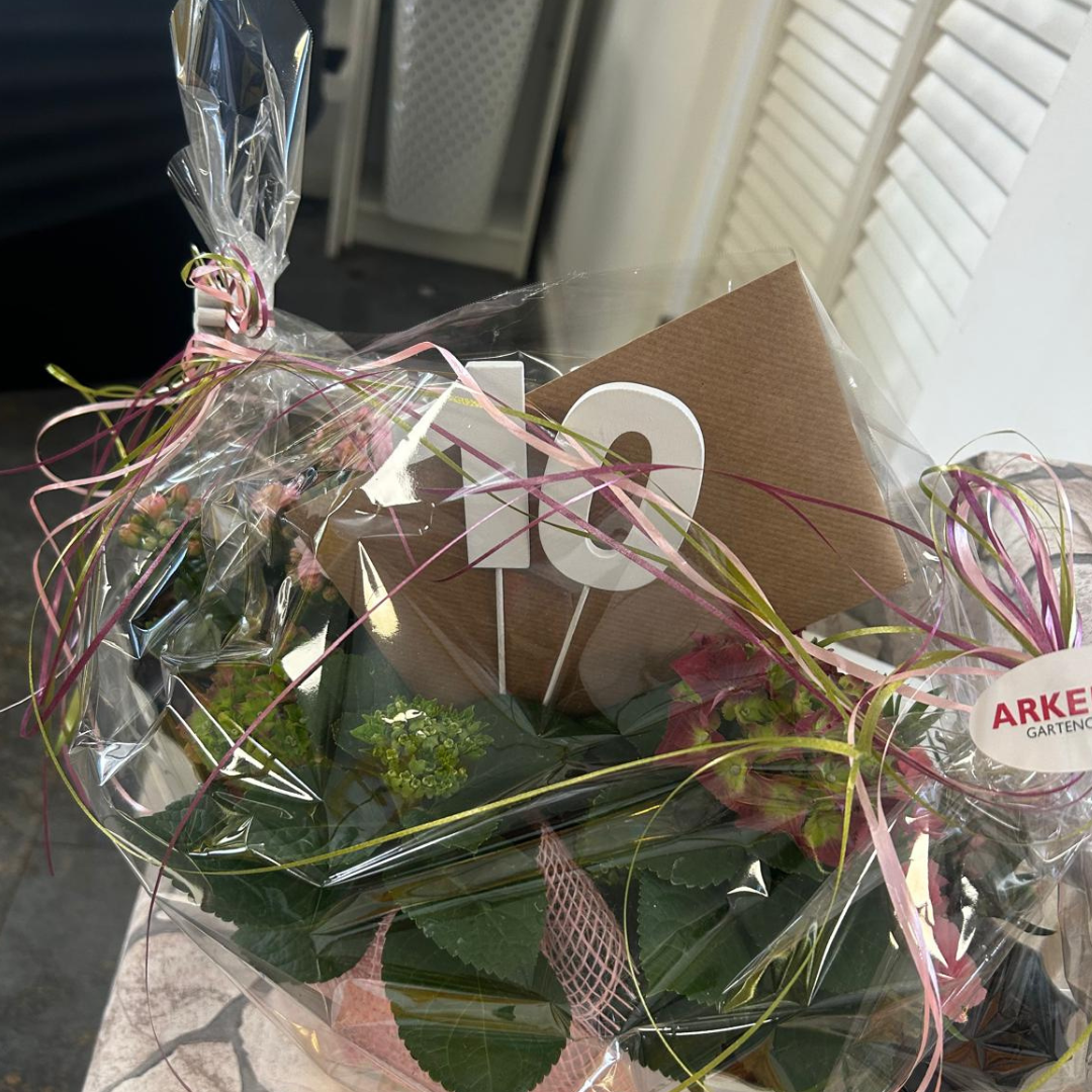 Gift bouquet wrapped in clear cellophane with pink and gold ribbons, a brown greeting card, and green foliage.