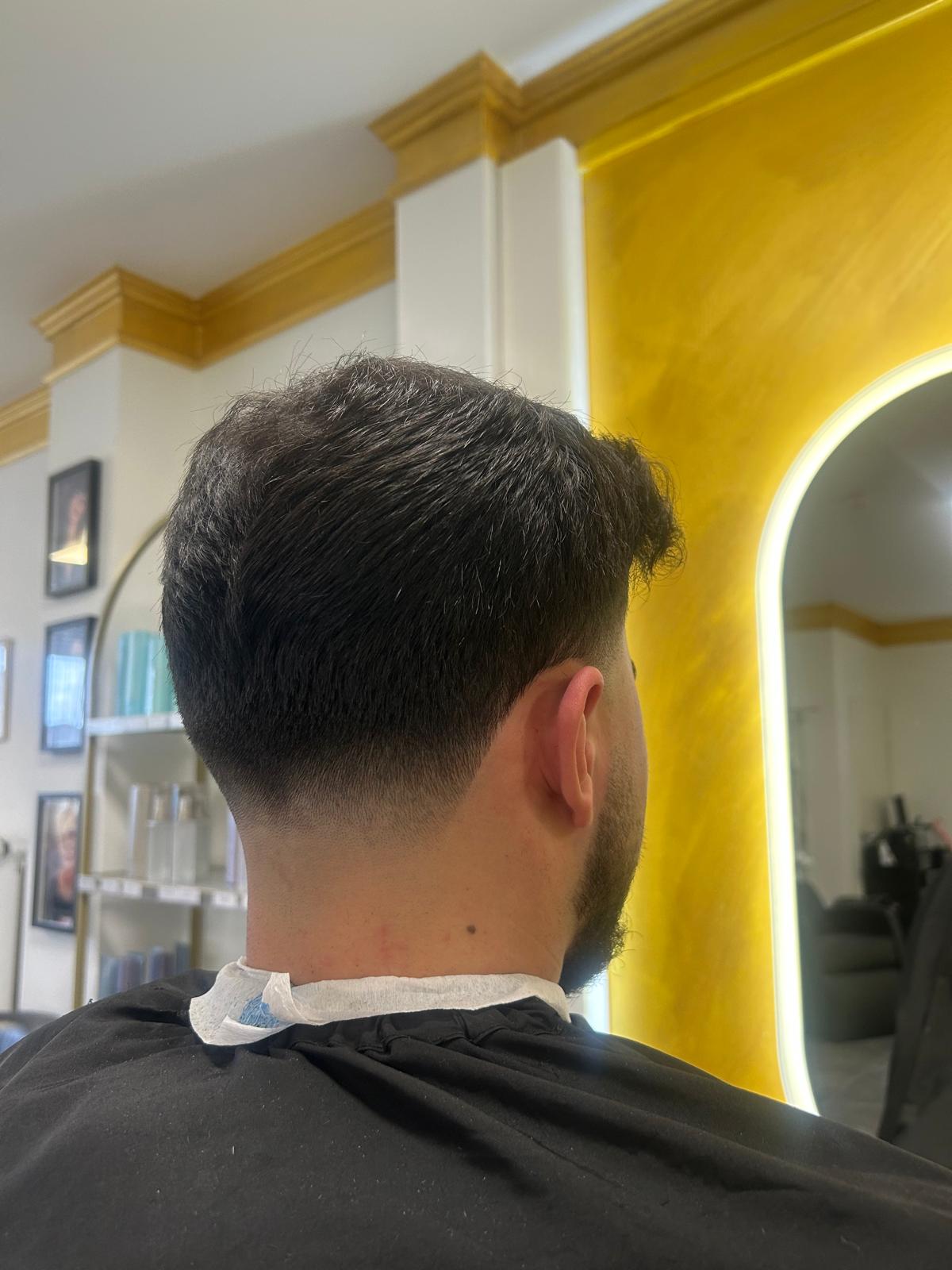Back view of a man with a low fade haircut, wearing a barber cape in a salon with a yellow wall and mirror nearby.